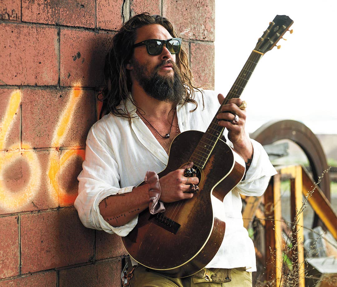 Jason Momoa in sunglasses and a white shirt plays guitar ي